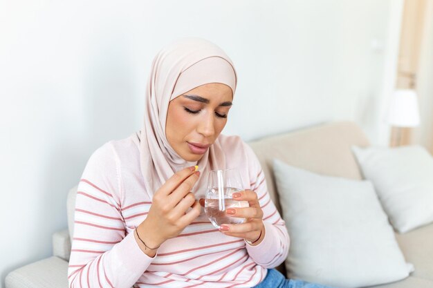 Konzept des Leidens unter schrecklichen Schmerzen Arabische traurige, verärgerte, unglückliche, unruhige, schwache Frau mit Hijab, die legere Kleidung trägt, sitzt auf einem Sofa und nimmt eine Pille