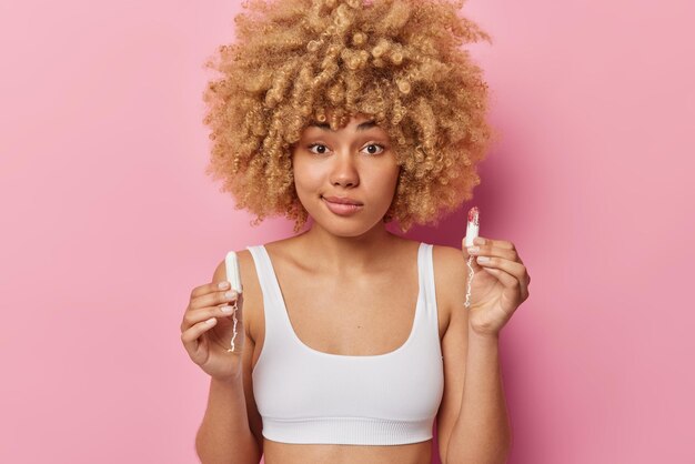 Konzept des Menstruationszyklus Junge zögerliche Frau mit lockigem Haar hält zwei Tampons für Perioden in weißem, kurz geschnittenem Oberteil, isoliert auf rosafarbenem Hintergrund, kümmert sich um die Intimhygiene und hat Blutungen