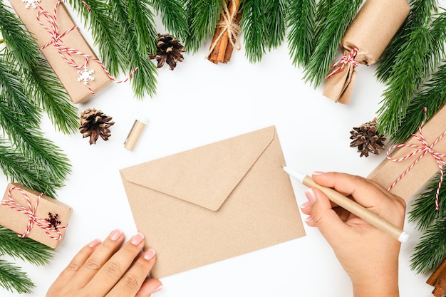 Konzept des schreibens des neujahrsbriefes an den weihnachtsmann, frau übergibt leeren handwerksumschlag mit kopienraum im rahmen von weihnachtsbaumzweigen, zapfen und geschenkboxen.