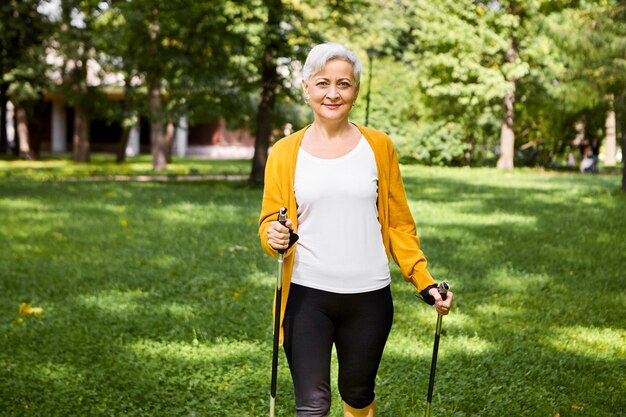 Konzept für Aktivität, Wohlbefinden, Sport und Ruhestand. Charming fit ältere Frau in stilvollen Radhosen und Strickjacke im Freien mit speziellen Stöcken posieren und skandinavischen Spaziergang im Park genießen