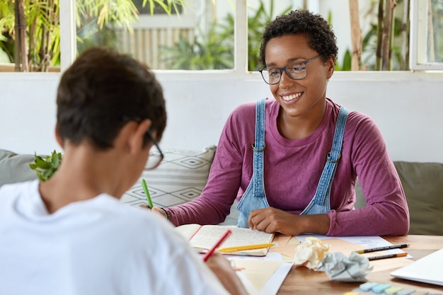 Konzept für Bildung, Beziehung und Zusammenarbeit. Zufriedene schwarze junge Frau mit lockigem Haar, trägt eine Brille zur Sehkorrektur, sieht Schüler positiv an, der versucht, Material zu verstehen