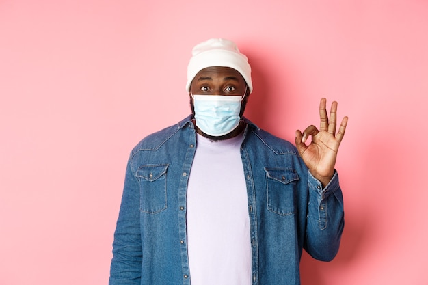 Konzept für Coronavirus, Lebensstil und soziale Distanzierung. Beeindruckter afroamerikanischer Mann mit Gesichtsmaske, der ein okayzeichen zeigt, wie und zustimmt, auf rosafarbenem Hintergrund stehend