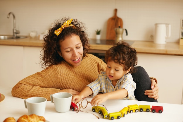 Konzept für Familie, Kinderbetreuung, Lernen, Entwicklung und Feinmotorik. Pflege der jungen hispanischen Frau, die Kaffee in der Küche trinkt, während hübscher kleiner Sohn, der neben ihr sitzt und mit Spielzeugbahn spielt