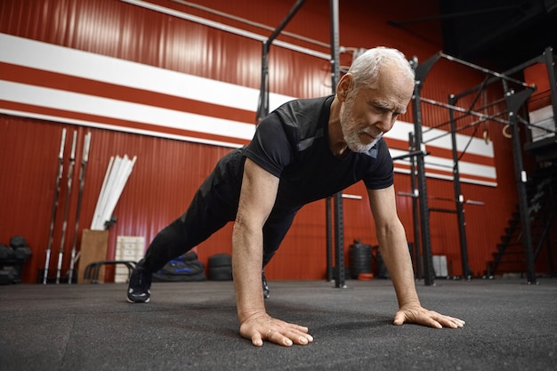 Konzept für Gesundheitswesen, Alter, Ruhestand und Rehabilitation. Muscle Fit 70 Jahre alter unrasierter Mann in Sportbekleidung, der Planke im Fitnessstudio tut. Ältere männliche Beplankung während des morgendlichen Trainings im Fitnesscenter