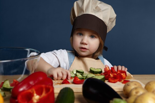 Konzept für Kinder, Lebensmittel, Ernährung und gesunden Lebensstil. Entzückendes freudiges 5 Jahre altes Baby in der Kochuniform, die verschiedene Gemüsesorten auf Kochbrett schneidet, während sie vegetarische Lasagne oder Suppe macht