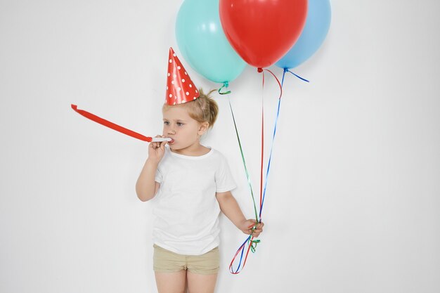 Konzept für Kindheit, Glück, Feier und Spaß. niedliches entzückendes kleines Kind, das Pfeife bläst, bunte Luftballons hält, sich glücklich fühlt, Geburtstag feiert, an weißer Wand posiert