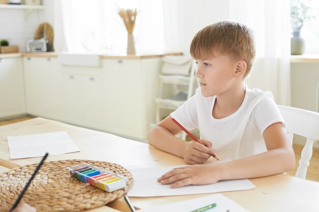 Konzept für Kreativität, Freizeit, Hobby, Kunst und Fantasie. Bild des nachdenklichen kaukasischen Schuljungen im weißen T-Shirt, das am Schreibtisch drinnen sitzt, nachdenklich schaut und überlegt, was mit Bleistift zu zeichnen ist