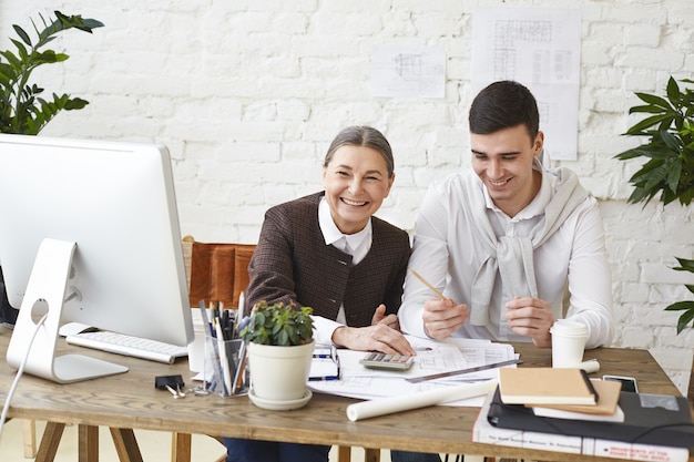 Konzept für Menschen, Arbeit, Teamwork und Zusammenarbeit. Glücklicher Chefingenieur mittleren Alters, der Spaß im Büro hat, während sie ihrem jungen männlichen Assistenten etwas erklärt, der mit PC und Papieren am Schreibtisch sitzt