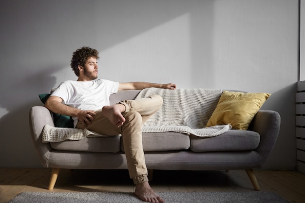 Konzept für Menschen, Lebensstil, Ruhe und Entspannung. Bild des gutaussehenden Kerls mit nackten Füßen, die drinnen ruhen, auf gemütlicher Couch sitzen und Augen schließen. Stilvoller junger Mann mit Stoppeln, die allein zu Hause entspannen