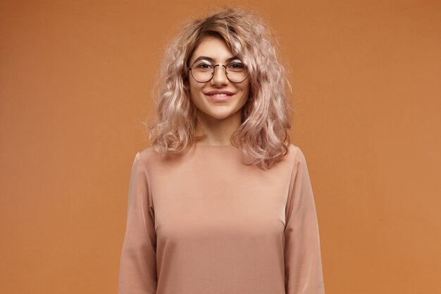 Konzept für Menschen, Lifestyle, Mode und Optik. Attraktives entzückendes europäisches Hipster-Mädchen mit voluminösem Haar und fröhlichem freundlichem Lächeln, das positive Gefühle ausdrückt