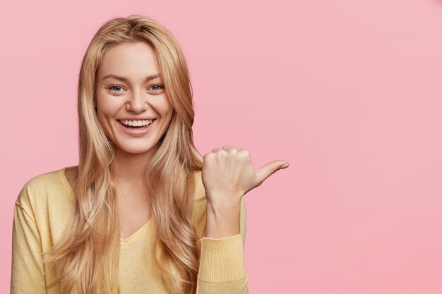 Konzept für Menschen, Werbung und Positivität. Glückliche junge Frau mit angenehmem Aussehen und Lächeln, zeigt mit dem Daumen zur Seite an leerem rosa Kopienraum für Ihren Werbetext oder Hörer an.