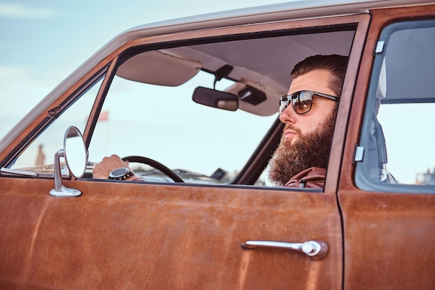 Konzept im Landhausstil. Bärtiger Mann mit Sonnenbrille in brauner Lederjacke, der ein Retro-Auto fährt.