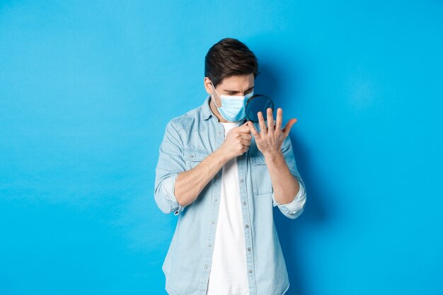 Konzept von Coronavirus, sozialer Distanzierung und Pandemie. Mann in medizinischer Maske, der seine Handfläche durch die Lupe betrachtet und auf blauem Hintergrund steht