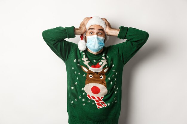 Konzept von Covid-19 und Weihnachtsferien. Aufgeregt und super glücklicher Mann in medizinischer Maske und Weihnachtsmütze, der erstaunt aussieht, weißer Hintergrund