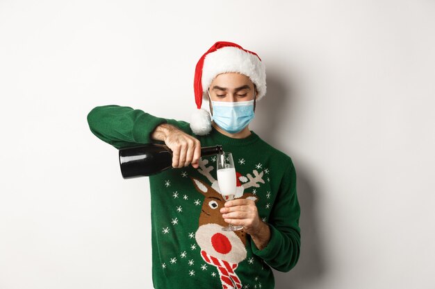 Konzept von Covid-19 und Weihnachtsferien. Fröhlicher Kerl in Gesichtsmaske und Weihnachtsmütze, Silvesterparty feiern und Glas Champagner gießen, weißer Hintergrund