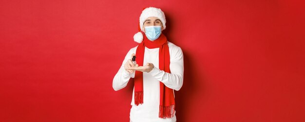 Konzept von Covid-19, Weihnachten und Feiertagen während der Pandemie. Glücklicher junger mann mit weihnachtsmütze und medizinischer maske, desinfiziert die hände mit antiseptikum und lächelt, steht auf rotem hintergrund.