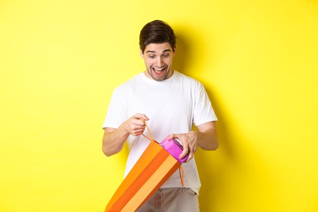 Konzept von feiertagen und feier. junger mann, der als herausnehmendes geschenk von der einkaufstasche überrascht schaut und über gelbem hintergrund steht.