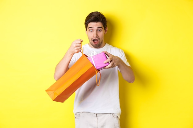 Konzept von Feiertagen und Feiern. Junger Mann, der überrascht aussieht, als Geschenk aus der Einkaufstasche herauszunehmen und auf gelbem Hintergrund steht.