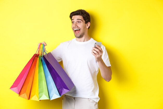 Konzept von Mobile Banking und Cashback. Glücklicher Mann, der erstaunt schaut, Einkaufstaschen und Smartphone, gelber Hintergrund hält.