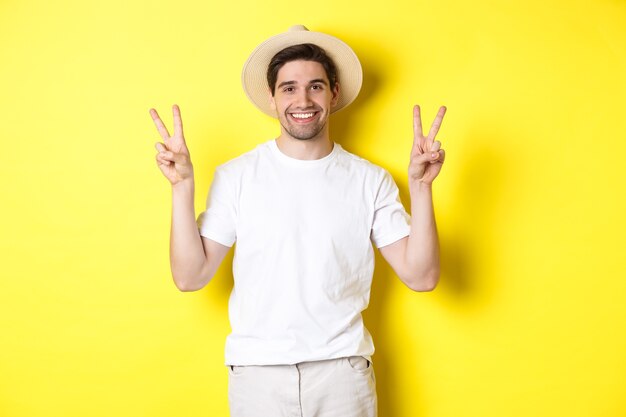 Konzept von Tourismus und Urlaub. Glücklicher männlicher Tourist, der für Foto mit Friedenszeichen aufwirft, aufgeregt lächelt, gegen gelben Hintergrund stehend.