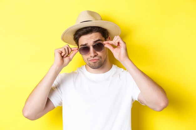 Konzept von Tourismus und Urlaub. Nahaufnahme des kühlen Touristen, der Feiertage auf Reise genießt, Sonnenbrille mit Strohhut, gelber Hintergrund tragend.