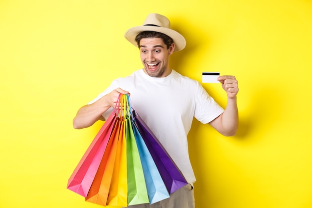 Konzept von Urlaub und Finanzen. Glücklicher Mannkäufer, der Einkaufstaschen zufrieden betrachtet, zeigt Kreditkarte, die gegen gelben Hintergrund steht.