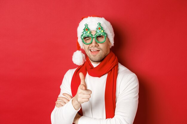 Konzept von Weihnachten, Winterferien und Feier. Nahaufnahme eines frechen jungen Mannes in Weihnachtsmütze und Partybrille, lächelnd und mit dem Finger auf die Kamera zeigend, über rotem Hintergrund stehend.