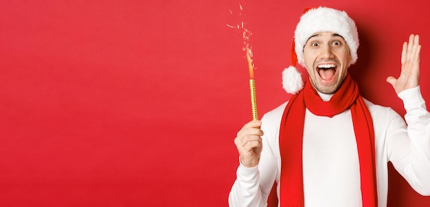 Konzept von Weihnachten, Winterferien und Feiern. Schöner Mann, der Neujahr feiert und Spaß hat, Wunderkerze hält und lächelt, Weihnachtsmütze trägt und über rotem Hintergrund steht.