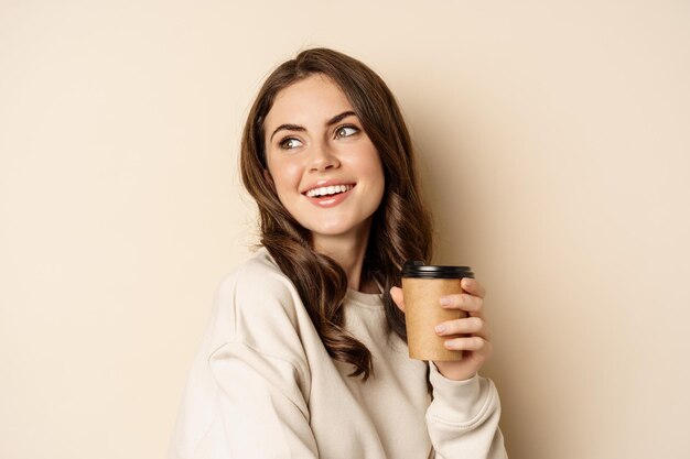 Konzept zum Mitnehmen und Café. Schöne weibliche lächelnde Frau, die eine Tasse Kaffee hält und vor beigem Hintergrund posiert. Platz kopieren