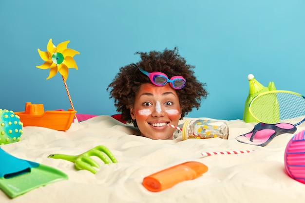 Kopf der jungen Frau mit Sonnenschutzcreme auf Gesicht umgeben von Strandaccessoires