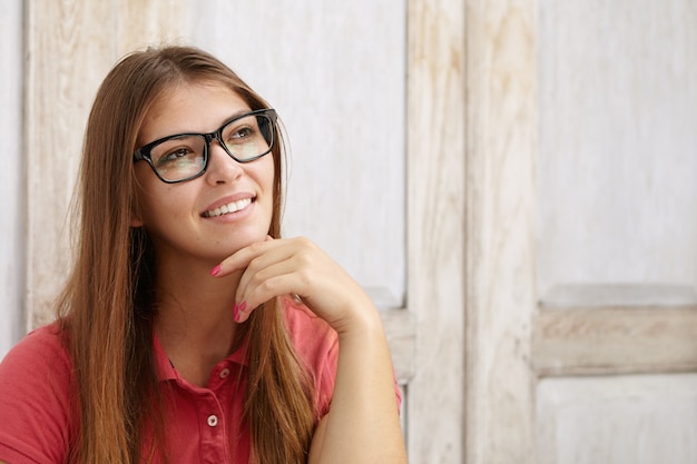 Kopfschuss der niedlichen jungen Frau, die Poloshirt und rechteckige Brille trägt, die Hand auf ihrem Kinn hält, während sie an etwas Angenehmes denken, lächelnd