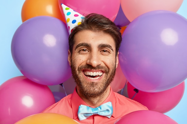 Kopfschuss des glücklichen europäischen Mannes trägt Papierkegelhut, rosa Hemd und Fliege, sieht positiv aus
