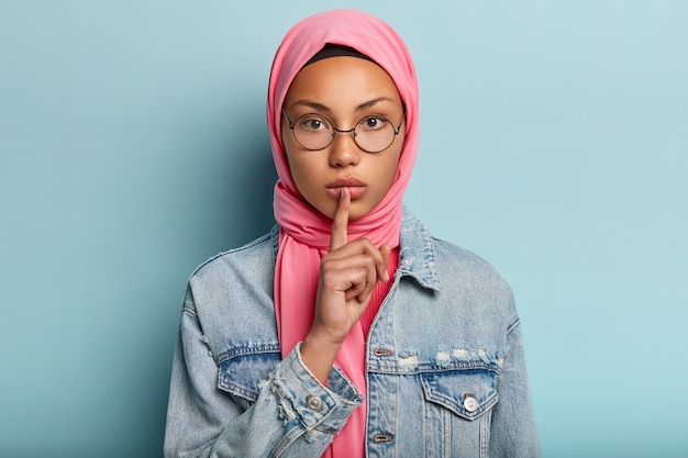 Kopfschuss einer ernsthaften Frau in rosa Schal, Jeansmantel, hält den Vorderfinger über den Lippen, bittet darum, keinen Lärm zu machen, macht eine leise Geste, hat islamische Überzeugungen, trägt eine runde optische Brille
