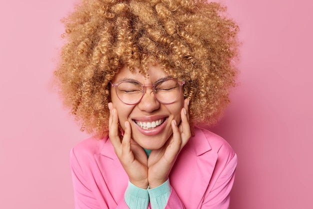 Kopfschuss einer glücklichen jungen Frau mit lockigem Haar hält die Augen geschlossen, die Hände auf den Wangen, grinst in die Kamera, trägt eine transparente Brille und formelle Jacke posiert vor rosa Hintergrund. Emotions-Konzept