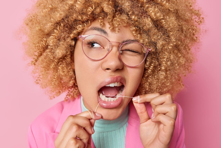 Kostenlose Foto kopfschuss einer jungen frau mit lockigem haar verwendet zahnseide, zwinkert mit den augen und kümmert sich um die konzentrierte mundhygiene. sie trägt eine transparente brille, elegante kleidung, isoliert auf rosafarbenem hintergrund
