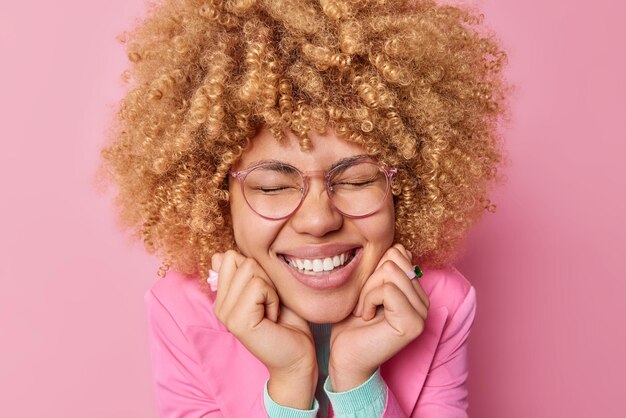 Kopfschuss einer optimistischen jungen Frau mit lockigen Haaren hält die Hände unter dem Kinn, lächelt zahnlos, ist gut gelaunt, trägt eine optische Brille, hört gute Nachrichten isoliert über rosa Hintergrund Fröhliche Emotionen