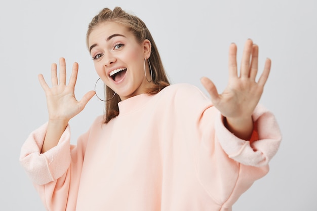 Kopfschuss eines attraktiven Lächelns mit offenem Mund, das ihre junge Frau mit weißen Zähnen zeigt, die in rosa langärmeligem Sweatshirt gekleidet ist und ihre Arme vor Freude ausstreckt. Glück und positive Emotionen.