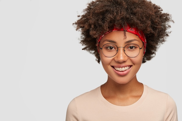 Kopfschuss eines glücklichen Hipster-Mädchens mit Afro-Haarschnitt, zufriedenem Gesichtsausdruck, entzückendem freundlichem Lächeln, Nostalgie, direktem Blick, weißer Wand, freiem Platz für Ihren Text