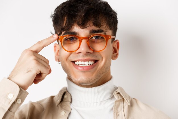 Kopfschuss eines lächelnden glücklichen Mannes, der einen neuen Brillenrahmen zeigt, auf die Brille zeigt und zufrieden aussieht und auf weißem Hintergrund steht.