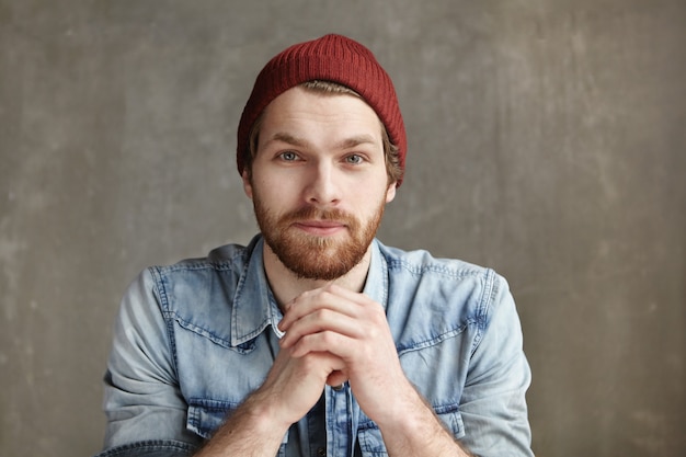 Kopfschuss eines modernen, gut aussehenden jungen europäischen Hipsters, der einen stilvollen Hut und ein Blue-Jeans-Hemd trägt, die Hände vor ihm gefaltet halten, nachdenklich und verträumt aussehen und an der Betonwand sitzen