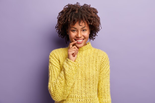 Kopfschuss von fröhlichen attraktiven weiblichen Blicken, posiert zum Fotografieren, lächelt breit, hält den Vorderfinger in der Nähe des Mundes, trägt einen gestrickten warmen Pullover, lacht über etwas Positives, isoliert auf Violett