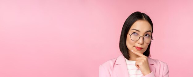 Koreanische Geschäftsfrau, die mit Brille denkt und nachdenklich in die Kamera schaut, um Entscheidungen zu treffen, die über rosa Hintergrund stehen