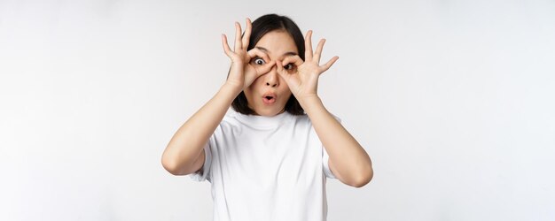 Koreanisches Mädchen der lustigen jungen asiatischen Frau, das Augenglasgeste macht, die die Kamera glücklich betrachtet, die über weißem Hintergrund steht