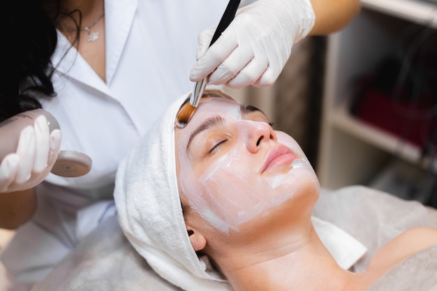 Kosmetikerin mit einem Pinsel trägt eine weiße Feuchtigkeitsmaske auf das Gesicht einer jungen Kundin in einem Spa-Schönheitssalon auf