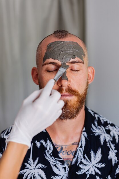 Kosmetikerin tragen Tonmaske mit Pinsel auf Gesicht des jungen gutaussehenden Mannes in der Schönheitsklinik auf