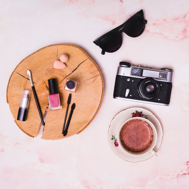 Kostenloses Foto kosmetikprodukte auf baumstumpf; kaffeetasse; weinlesekamera und -sonnenbrillen auf rosa strukturiertem hintergrund