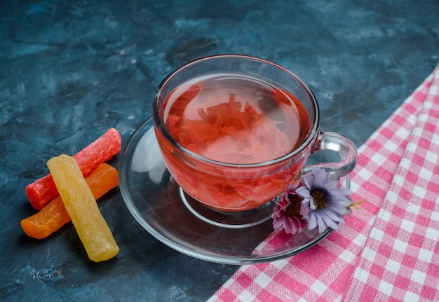 Kräutertee mit Süßigkeiten, Blumen in einer Tasse auf Blau und Geschirrtuch, hohe Winkelansicht.