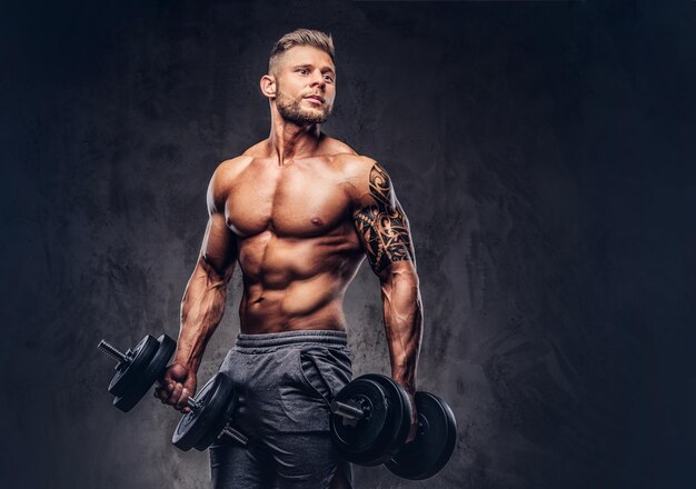 Kraftvoller, stylischer Bodybuilder mit Tätowierung auf dem Arm, der die Übungen mit Hanteln macht. Getrennt auf einem dunklen Hintergrund.
