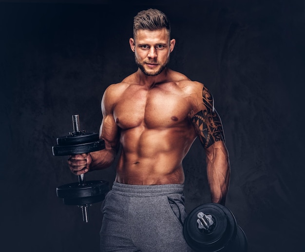 Kraftvoller, stylischer Bodybuilder mit Tattoo auf dem Arm, posiert mit Hanteln in einem Studio. Getrennt auf einem dunklen Hintergrund.