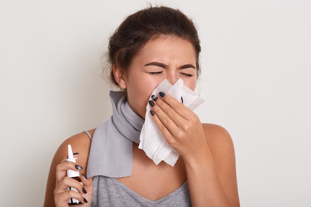 kranke allergische Frau, die laufende Nase putzt, Grippe hat oder sich erkältet, im Taschentuch niest, mit geschlossenen Augen auf Weiß isoliert posiert, Nasenspray in der Hand hält.